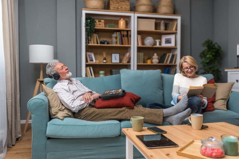 Mann og kvinne i sofa som lytter til DAISY spiller stratus 2. Mann har på hodetelefoner og kvinnen leser en bok.
